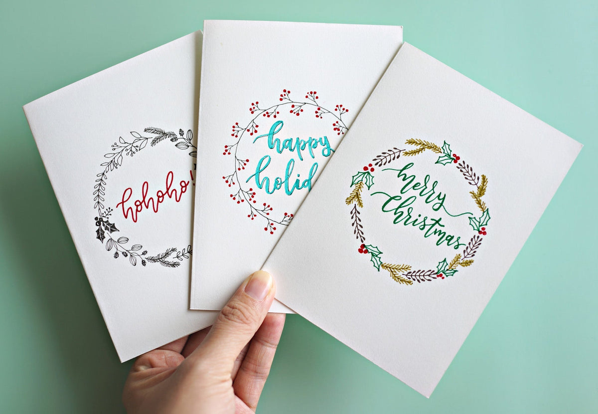 A hand holding three white Christmas cards with hand-lettered wreath designs, against a mint green background.