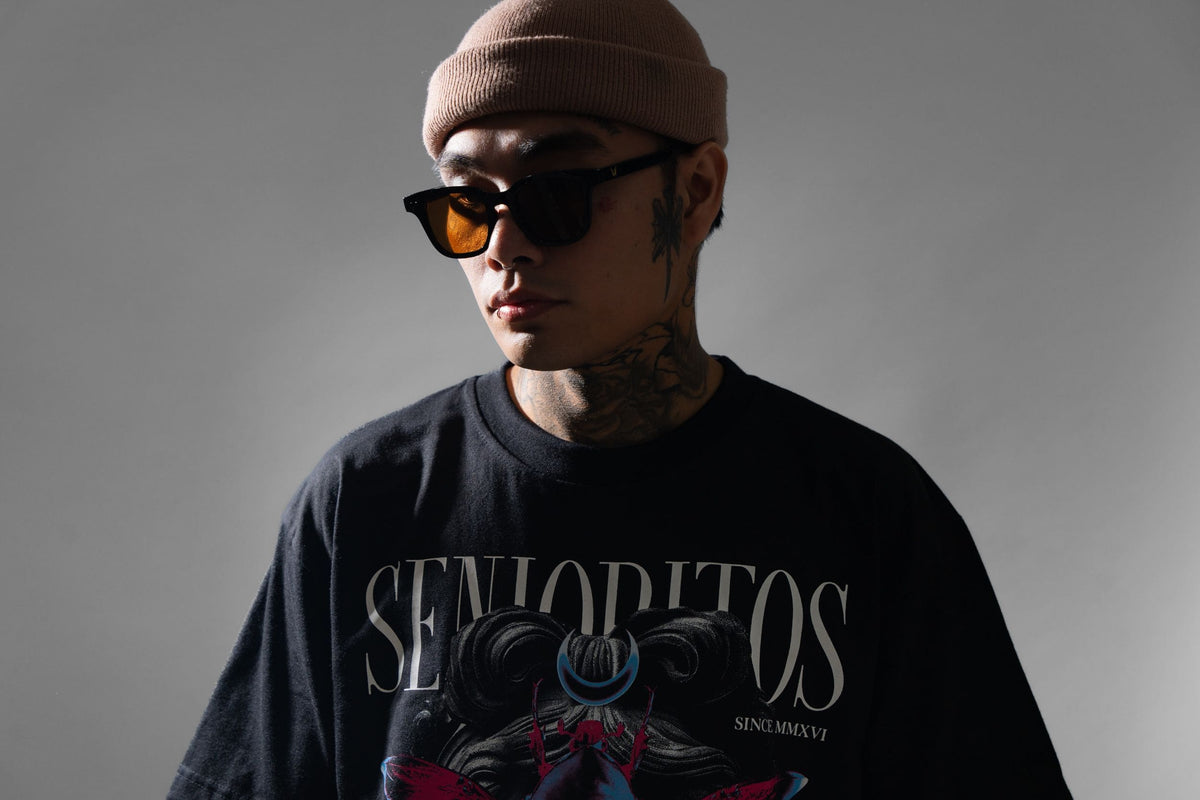 Studio portrait of a man in a beige beanie, dark sunglasses with orange lenses, and a black graphic T-shirt reading "SEÑORITOS," against a grey background.